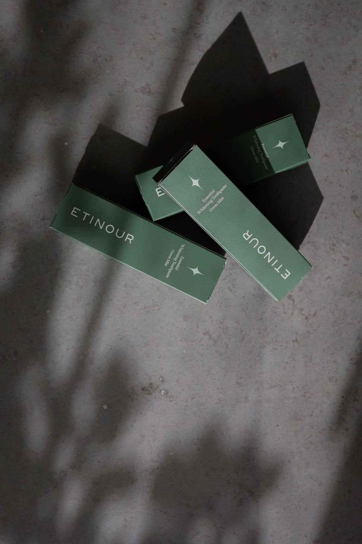 Three Etinour Essential Whitening Toothpaste outer green packaging in a row, on a grey stone surface and shadows on top